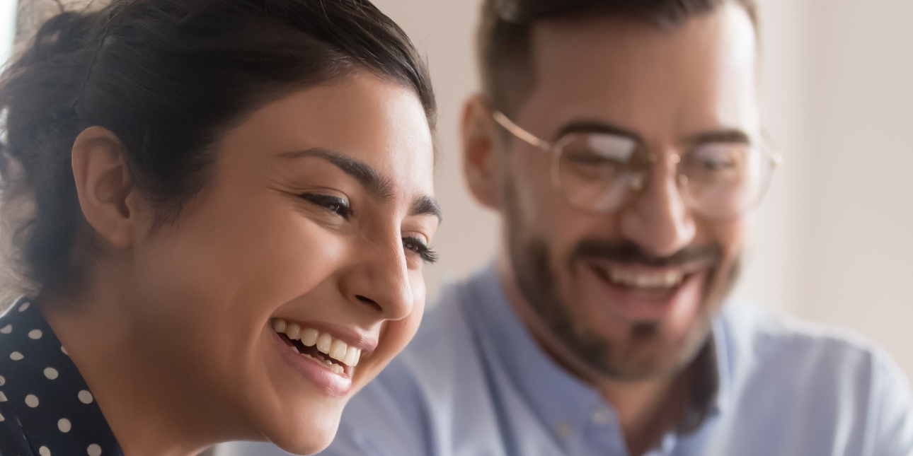 An Asian woman with short dark hair wearing a polka dot shirt laughs with a white male colleague who is wearing glasses, had a beard and has a blue shirt